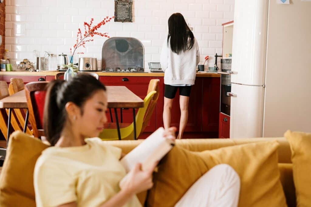 a woman reading a book on a sofa, another woman standing in the kitchen