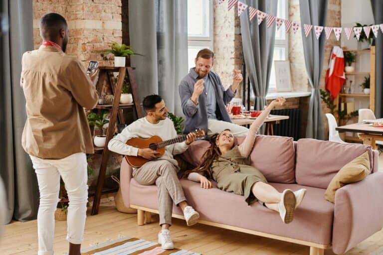 A group of four friends enjoying a relaxed gathering in a stylish living room, with one playing the guitar, another dancing, and a woman lounging on a pink couch.