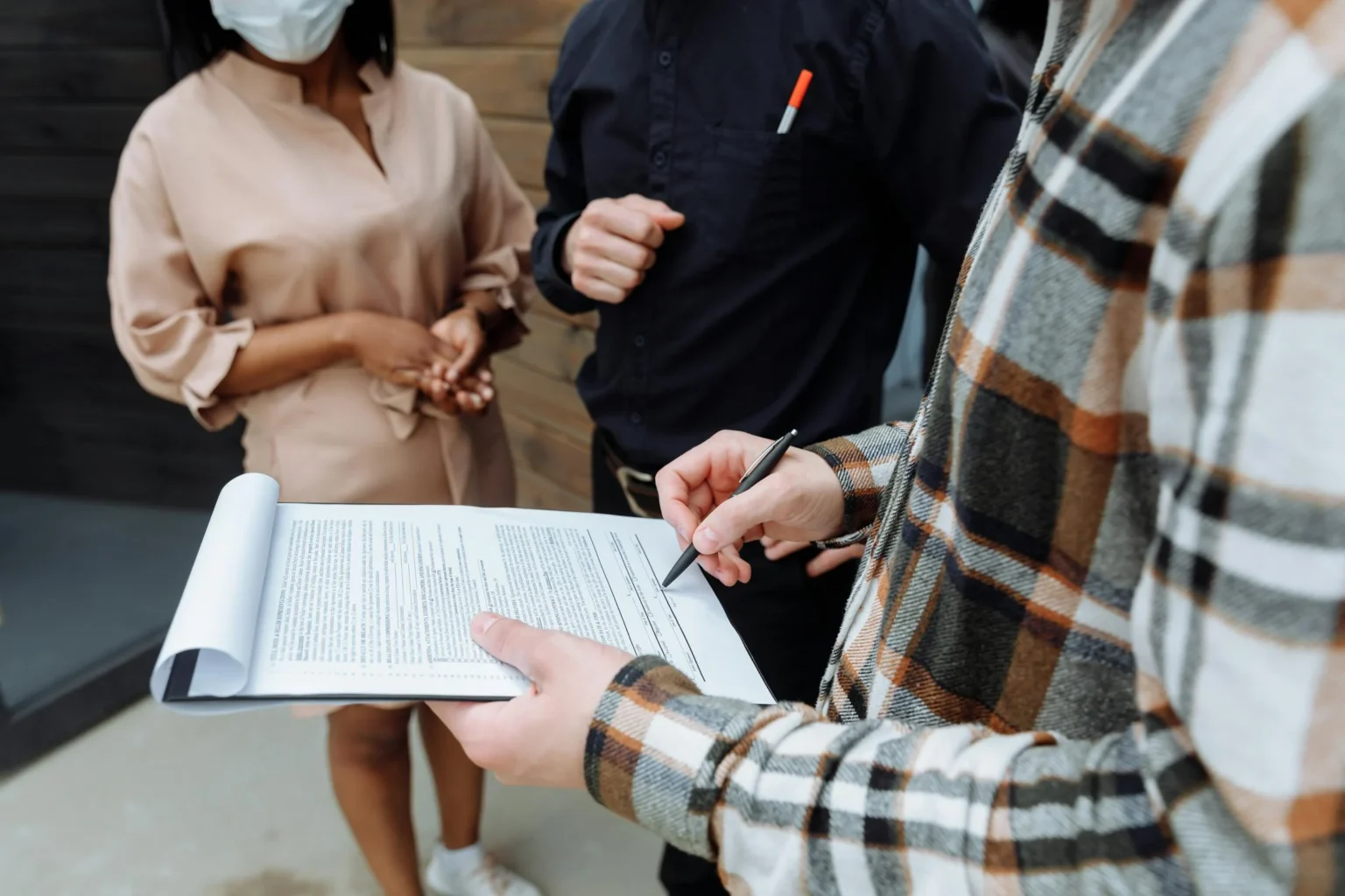 Close-up of a person signing a document on a clipboard while two professionals stand in the background, discussing terms