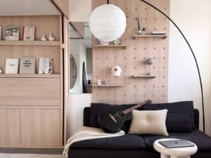 Stylish small living space with pegboard wall, bookshelves, a black couch, and personal items like a guitar and decor.