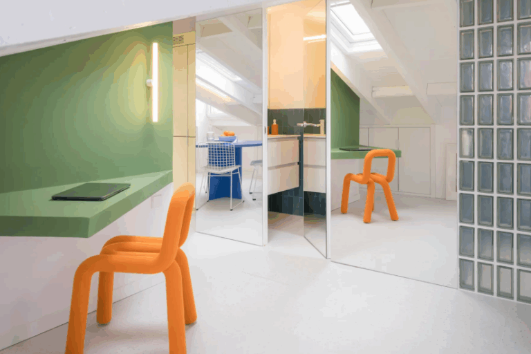 Small attic apartment with green wall-mounted desk, orange chair, mirrored closet doors, and built-in sloped ceiling storage.