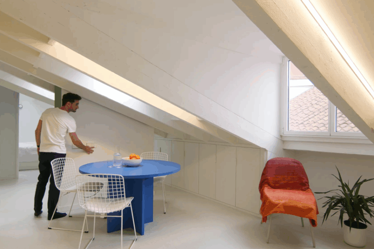 Man in a white shirt beside a blue dining table and white wire chairs under a sloped ceiling with custom storage and a large window.