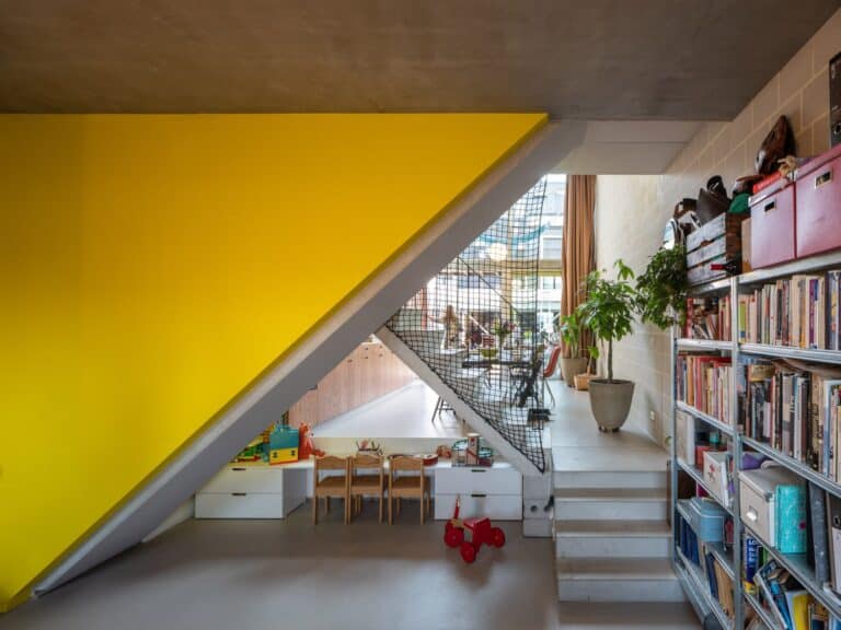 Modern interior space featuring a bright yellow wall under a staircase, a small children’s play area, a netted safety barrier, and a cozy communal living area with bookshelves, plants, and natural lighting.