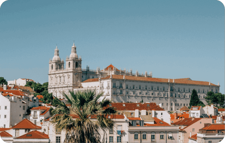 Sunlit view of historic Lisbon buildings with palm tree and church under clear blue sky