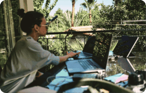 Person working with multiple laptops outdoors on a sunny balcony, surrounded by lush greenery