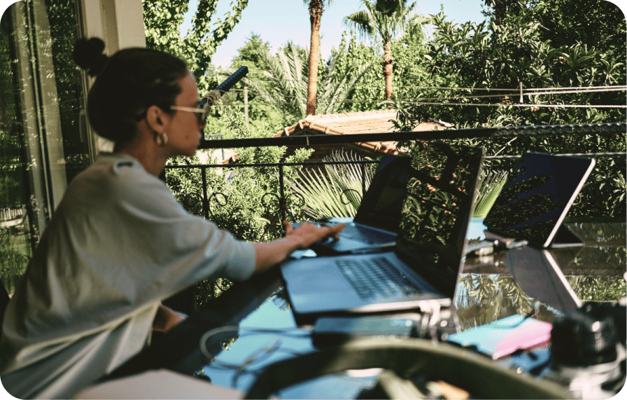 Person working with multiple laptops outdoors on a sunny balcony, surrounded by lush greenery