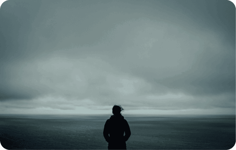 Person standing alone by the ocean under a cloudy, moody sky, symbolizing loneliness and isolation