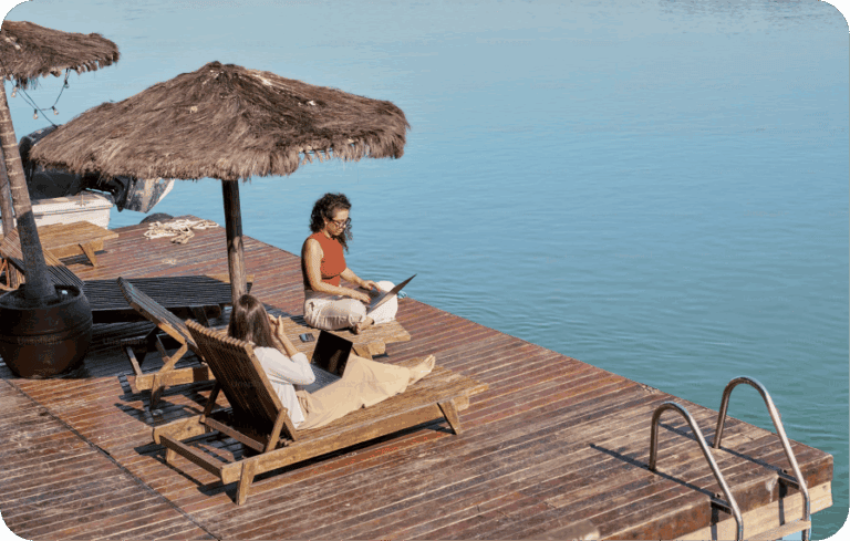 Two women working on laptops on wooden deck beside water under straw sun umbrellas